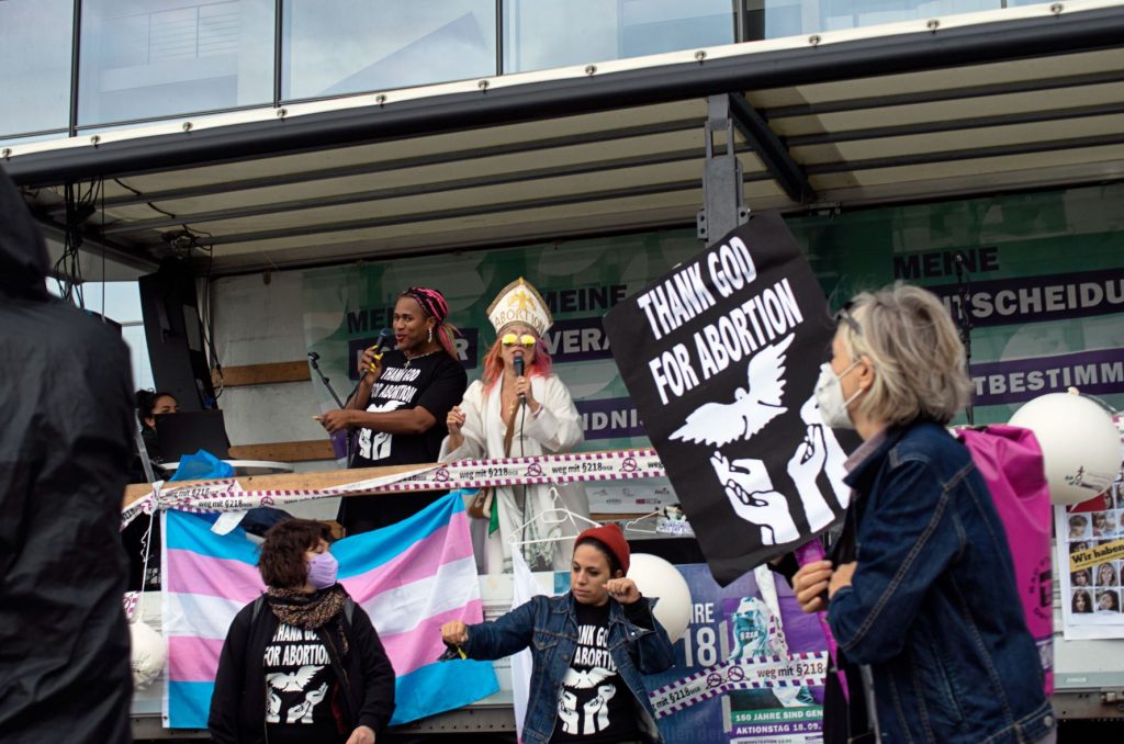 Zwei Personen stehen auf einer Bühne und halten einen Redebeitrag auf einer Pro-Choice-Kundgebung. Im Forderung ist ein Schild mit der Aufschrift "Thank God for Abortion" sichtbar.