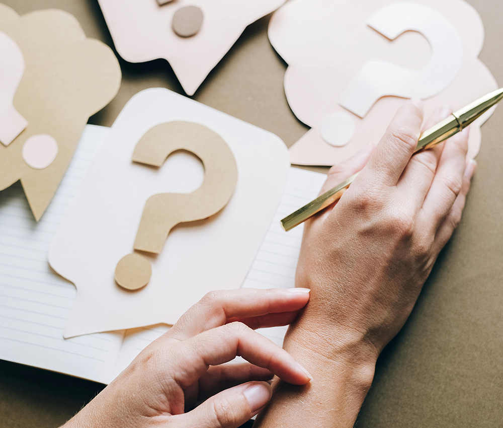 Die Hände einer Person ruhen auf einem Tisch, in der einen Hand hält die Personeinen Stift, auf dem Tisch liegen Bilder mit großen Fragezeichen darauf.