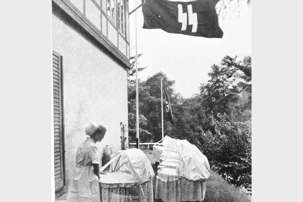 Eine Person mit Krankenschwester-Uniform steht mit Wiegen unter einer SS-Flagge