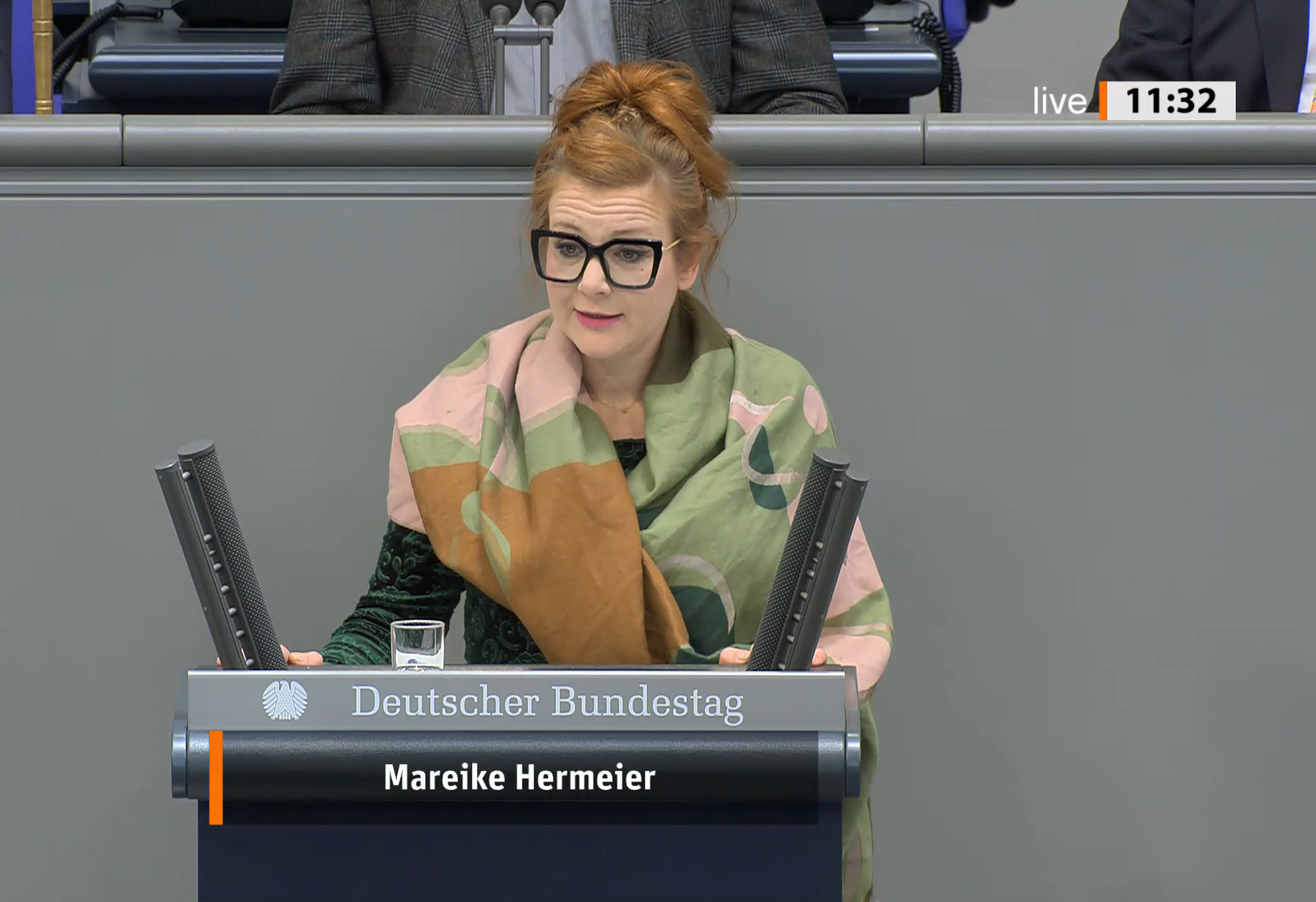 Politikerin Mareike Hermeier am Rednerpult im Plenarsaal des Bundestags. Hermeier ist weiß, sie trägt ihr rötliches Haar zu einem hochsitzenden Dutt gebunden. Hermeier trägt eine schwarz gerahmte Brille undd ein schwarzes Oberteil. Um ihre Schultern ist ein breiter grüner Schal geschlungen.