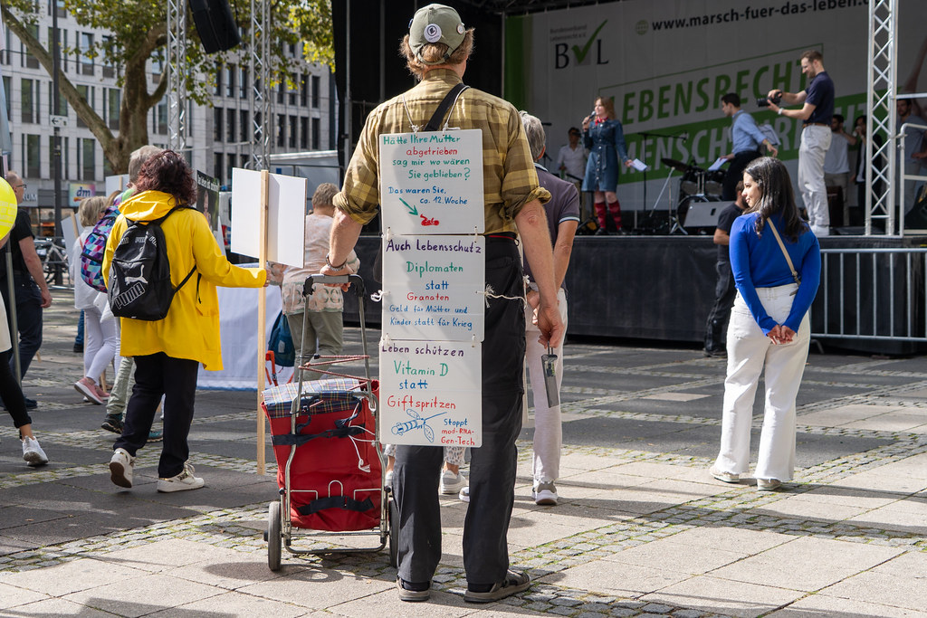 Person mit Schild auf dem Rücken bei einer Kundgebung auf einem Platz vor einer Bühne mit mehreren Menschen