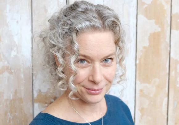 Nahaufnahme von Alys Weinbaum. Sie hat grau-blonde Locken und trägt einen blauen Pullover mit rundem Ausschnitt sowie eine Halskette.