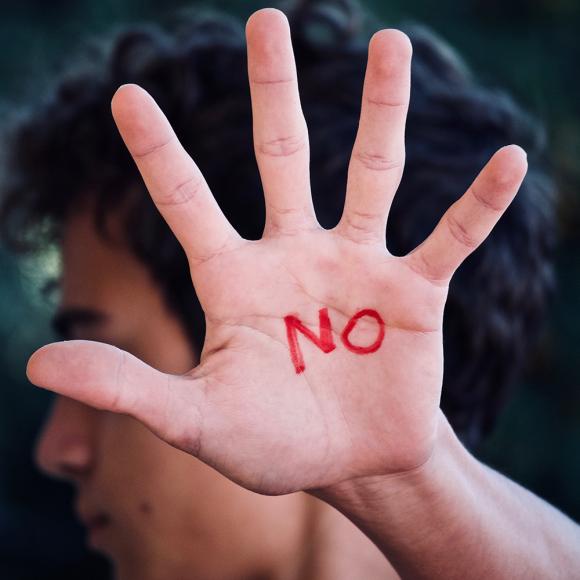Im Vordergrund ist die Hand einer Person zu sehen, Finger sind gespreizt, in der Handfläche steht das Wort "NO", im Hintergrund ist unscharf das Profil der Person zu sehen.