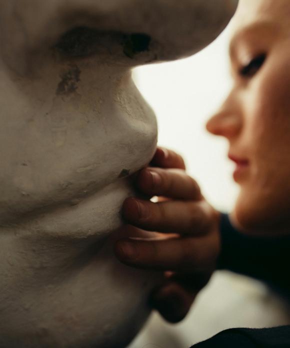 Das Gesicht einer weiblich gelesenen Person ist teilweise im Profil zu sehen, ihre Hand berührt die Lippen einer großen Statue oder Büste, von der nur ein Ausschnitt des Gesichts zu erkennen ist.