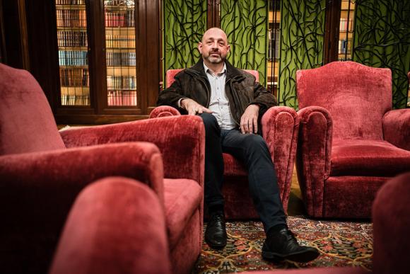 Yves Moreaux sitzt in einem roten Samtsessel und blickt geradewegs in die Kamera. Er trägt ein helles Hemd, eine braune Lederjacke und eine schwarze Jeans. Daneben stehen weitere rote Samstsessel und im Hintergrund sind Bücherregale und künstvolle Fenster zu erkennen.