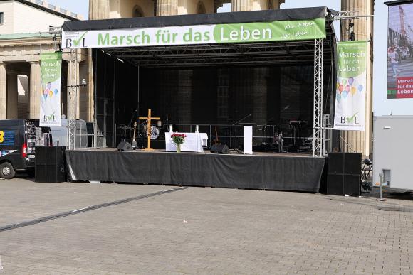 Eine leere Bühne. Oben unter der Decke ein Banner mit der Aufschrift "Marsch für das Leben". Auf der Bühne ein Rednerpult und ein großes Holzkreuz.
