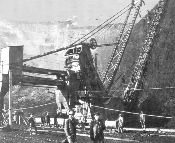 Ein historisches Schwarz-Weiß-Foto zeigt eine große, komplexe Bergbaumaschine in einem Steinbruch oder Tagebau. Mehrere Arbeiter in Arbeitskleidung und Mützen stehen im Vorder- und Hintergrund verteilt, einige nahe kleiner Loren auf Schienen. Die Maschine besteht aus hohen Stahlträgern, Seilen und einem großen Schaufel- oder Fördermechanismus, der sich entlang einer steilen Felswand erstreckt. Die Szene vermittelt frühe industrielle Schwerarbeit.