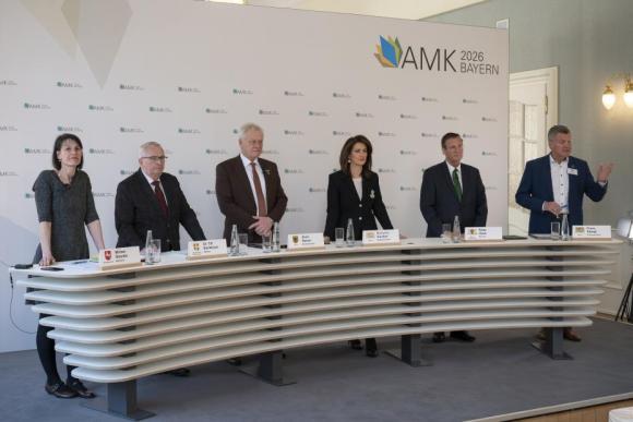 Fünf Personen stehen hinter einem langen, geschwungenen Pult mit Namensschildern und Wassergläsern, im Hintergrund eine Wand mit dem Schriftzug 'AMK 2026 BAYERN'