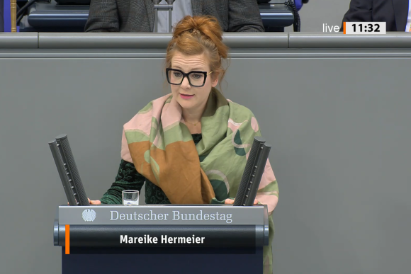 Politikerin Mareike Hermeier am Rednerpult im Plenarsaal des Bundestags. Hermeier ist weiß, sie trägt ihr rötliches Haar zu einem hochsitzenden Dutt gebunden. Hermeier trägt eine schwarz gerahmte Brille undd ein schwarzes Oberteil. Um ihre Schultern ist ein breiter grüner Schal geschlungen.